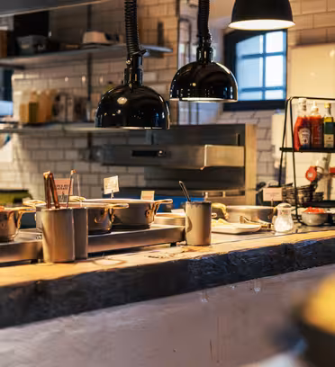 Et hyggeligt restaurantkøkken med forskellige gryder og pander på en stenbord, under sorte pendellamper. Hylder holder krydderier og forsyninger mod en hvid flisebelagt væg. Et lille vindue lukker naturligt lys ind.