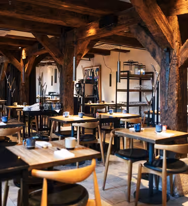 Et rustikt restaurantinteriør med træbjælker og søjler. Værelset har træborde og -stole, hvert sæt med stearinlys og service. Hylder med glasvarer langs bagvæggen, og belysningen er varm og imødekommende.