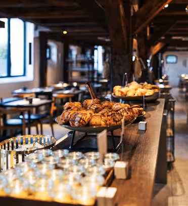 En hyggelig café med træbjælker har et langbord med en række forskellige kager, herunder croissanter. Glas og krydderier er pænt arrangeret i nærheden. Lokalet er blødt oplyst med flere borde og stole i baggrunden.