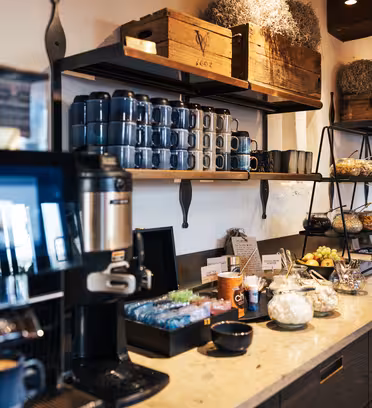 En moderne kaffebar med kaffemaskine, engangskopper, teposer og en række forskellige krus på hylderne. Disken har krukker med sukker, en skål frugt, kager og indretning. Stemningen er varm og indbydende.