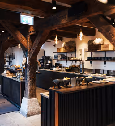 Rustikt restaurantinteriør med synlige træbjælker og industriel belysning. Buffetborde rummer tallerkener og fade, med hylder fyldt med krukker og indretning. En hyggelig, moderne atmosfære med neutrale farver og jordagtige accenter.