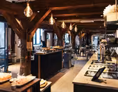 Et rummeligt, varmt oplyst restaurantinteriør med træbjælker og en buffetopstilling. Rustik indretning suppleres med hængende pærer. Der er arrangeret borde med mad, hvilket skaber en indbydende spisestemning.