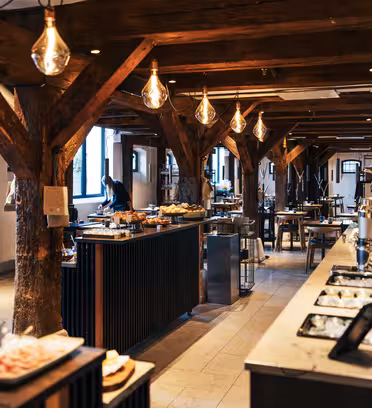 Et rummeligt, varmt oplyst restaurantinteriør med træbjælker og en buffetopstilling. Rustik indretning suppleres med hængende pærer. Der er arrangeret borde med mad, hvilket skaber en indbydende spisestemning.