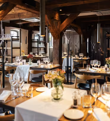 Et hyggeligt restaurantinteriør med træbjælker og borde dækket med hvide duge, vinglas og blomsterarrangementer. Blødt lys skaber en varm stemning. Hylder med glasvarer er synlige i baggrunden.