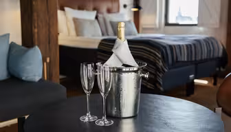 Et hyggeligt soveværelse har en seng med stribet sengetøj og puder. I forgrunden rummer et sort bord en sølvisspand med en indpakket champagneflaske og to tomme fløjteglas. Ruskindspuder hviler på en nærliggende sofa.
