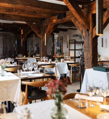 Rustikt restaurantinteriør med træbjælker og borde til spisning. Hvide duge, pænt opstillede stole, poleret glas og midterstykker af friske blomster skaber en indbydende atmosfære. Blød belysning tilføjer den varme stemning.