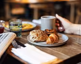 Et bord med morgenmadsartikler, herunder en rulle med frø, croissant og kager på en tallerken. En person holder et krus. Også synlige er to glas med drinks og en foldet avis ved siden af bestik på en serviet.