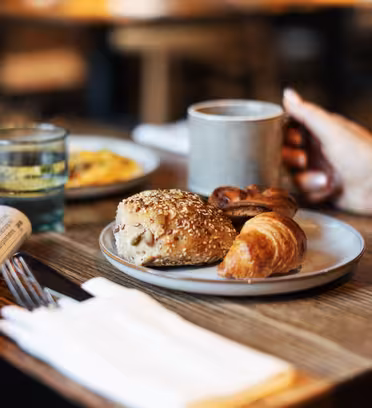 Et bord med morgenmadsartikler, herunder en rulle med frø, croissant og kager på en tallerken. En person holder et krus. Også synlige er to glas med drinks og en foldet avis ved siden af bestik på en serviet.