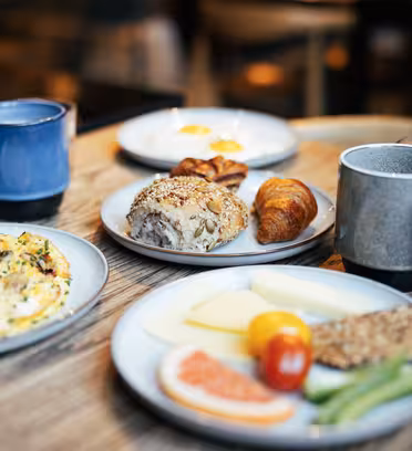 Et træbord med morgenmadsartikler: en omelet drysset med urter, diverse brød, osteskiver, en tomat, agurk, grapefrugtskive, kaffekrus og glas vand. Baggrunden er blødt sløret.