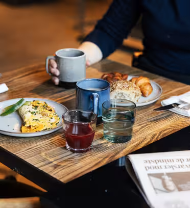 En person, der sidder ved et træbord med en tallerken med omelet, croissanter og en rundstykke. De holder et krus og har to glas juice og vand i nærheden. En foldet avis ligger på bordet sammen med to servietindpakkede redskaber.