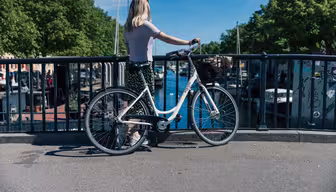 En kvinde med lyst hår står ved siden af en cykel og kigger over et brorækværk på en fredfyldt kanal. Bådene ligger til kaj i vandet, og træer kanter baggrunden under en klar blå himmel.