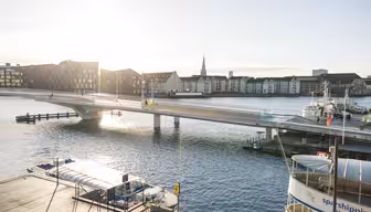 En solbelyst scene ved vandet med en moderne gangbro, der strækker sig over en rolig flod. Bygninger langs vandet, og et par både ligger til kaj i nærheden. Himlen er klar og solen kaster refleksioner på vandet.