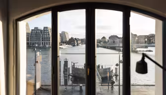 Udsigt gennem et buet vindue, der viser en scene ved vandet med moderne og traditionelle bygninger. En rolig flod afspejler den overskyede himmel, mens en strandpromenade fuld af mennesker bringer det rolige bybillede til live.