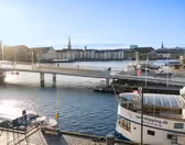 Udsigt over en soloplyst scene ved vandet med en bro, der krydser en kanal. Cyklister er på broen, og flere både ligger fortøjet i nærheden. I baggrunden ses historiske bygninger og kirkespir under en klar blå himmel.
