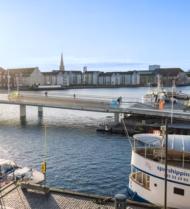 Udsigt over en soloplyst scene ved vandet med en bro, der krydser en kanal. Cyklister er på broen, og flere både ligger fortøjet i nærheden. I baggrunden ses historiske bygninger og kirkespir under en klar blå himmel.