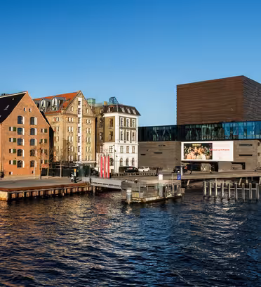 Udsigt ved vandet med en række historiske murstensbygninger og en moderne glasstruktur med et fladt tag, der strækker sig over vandet. Bådene ligger til kaj langs træmolerne under en klar blå himmel.
