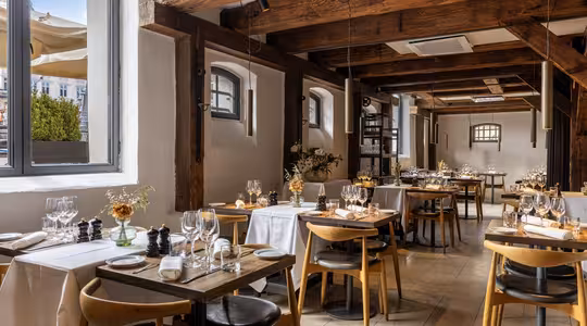 En hyggelig, rustik restaurant med træbjælker, hvide vægge og store vinduer. Bordene er dækket med hvidt sengetøj, glas og elegant service, hvilket skaber en indbydende, varm atmosfære.