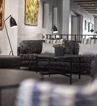 En hyggelig lounge med moderne sofaer med mønstrede hynder og sorte sofaborde. På bordene stilles dekorative plantekrukker. Rummet er prydet med farverige kunstværker på de hvide vægge og oplyst af en række loftslamper.