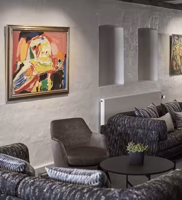 En hyggelig lounge med mørke sofaer, en grå lænestol og runde borde. Farverige abstrakte malerier pryder de lysegrå vægge med smalle vinduer, og moderne gulvlamper giver lys. Små grønne planter er placeret på bordene til indretning.