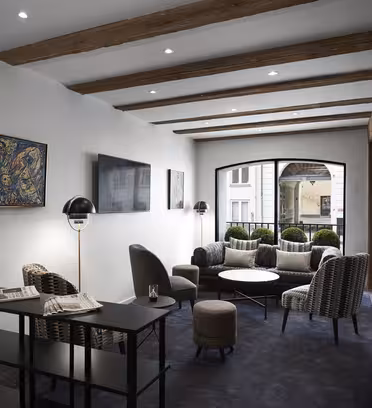 Et moderne loungeområde med træbjælker i loftet, med stole, små runde borde og en hylde med aviser. Kunstværker dekorerer væggene. Store vinduespartier giver naturligt lys og afslører udsigten til en gårdhave med grønne områder.