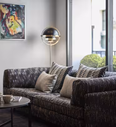 En moderne stue med en mørk, struktureret sofa prydet med mønstrede puder. Et rundt sofabord rummer krus og en skål. En gulvlampe står i nærheden. Store vinduespartier afslører en terrasse med grønne planter og bygningsfacader. Et farverigt maleri er på væggen.
