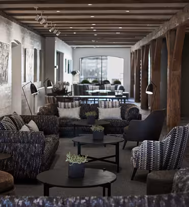 En hyggelig, moderne lounge med synlige træbjælker og en række polstrede stole og sofaer. Cirkulære sofaborde med små planter er placeret foran siddepladserne. Blød overheadbelysning skaber en varm stemning med kunst på væggene.