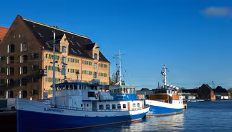 To blå og hvide både lagde til i nærheden af en murstensbygning med grønne skodder under en klar blå himmel. Bygningen har et stejlt tag med kvistvinduer. Sollys oplyser scenen og kaster refleksioner på vandet.