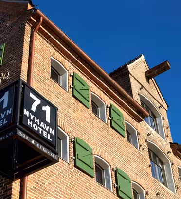 Murstensbygning under klar blå himmel med et hjørneskilt, hvor der står "71 NYHAVN HOTEL." Bygningen har grønne vinduesskodder og en markant, traditionel arkitektonisk stil.