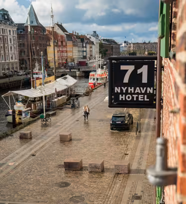 Udsigt over en brostensbelagt gade langs en kanal med farverige bygninger og fortøjede både. Et skilt med teksten "71 Nyhavn Hotel" er synligt på en murstensbygning. Et par går forbi cykler på en solskinsdag med luftige skyer på himlen.