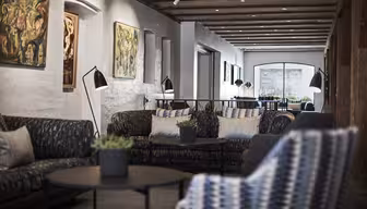 Moderne lounge med mønstrede sofaer og runde borde, dekoreret med potteplanter og kunst på hvide murstensvægge. Loftsbjælker og sporbelysning bidrager til den hyggelige og moderne atmosfære. Stort vindue yderst.