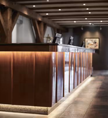 Et moderne hotelreceptionsområde med en trædisk, oplyst nedefra. Skranken har computere på toppen, og rummet har synlige træbjælker og malerier på væggene. Belysningen er blød og varm, hvilket skaber en indbydende atmosfære.