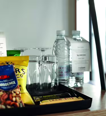 En hotelværelsesbakke med en flaske rødvin, pakker med snacks, to vandflasker og fire glas. Der er informationskort bag varerne. Bakken placeres på en træoverflade nær store vinduer.