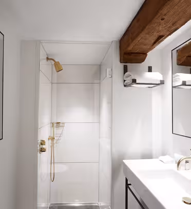 Minimalistisk badeværelse med hvide vægge og loft. Har brusekabine med messingarmaturer, moderne hvid vask med messinghane og træbjælker over hovedet. To spejle hænger over vasken, hver med en hylde med håndklæder.