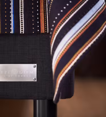 Nærbillede af en stol med et dekorativt stribet stof draperet over ryglænet. En sølvplade på stolen lyder "Sveriges Carpe Diem-senge." Scenen er varmt oplyst, hvilket fremhæver stolens slanke design.