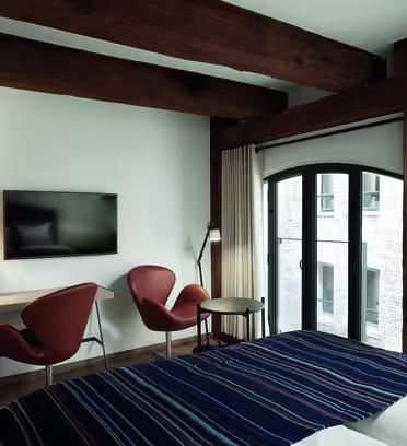 Hotelværelse med træbjælker, en seng med et stribet tæppe, to brune stole, et lille bord og et væghængt tv. En gulvlampe står nær vinduet, som har et buet design og udsigt til murstensbygninger udenfor.