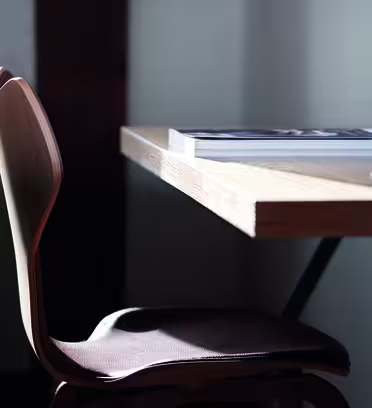 En træstol er placeret ved siden af et lyst træbord. Et lukket magasin hviler på bordet under blød belysning. Scenen formidler et minimalistisk og fredfyldt arbejdsrum.