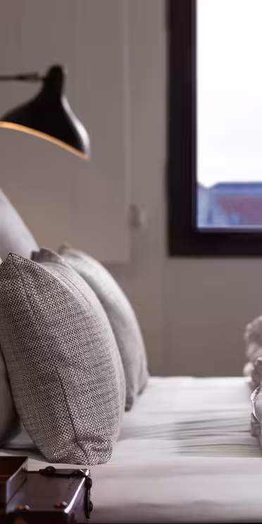 Et hyggeligt soveværelse med en pæn opredt seng med hvidt sengetøj og grå puder. To sorte væglamper giver belysning på den polstrede sengegavl. Et vindue giver en fjern udsigt over et bybillede med et spir i baggrunden.
