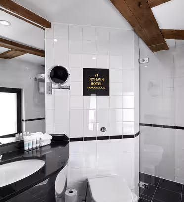Moderne hotelbadeværelse med sorte og hvide fliser. En elegant sort bordplade har en vask, toiletartikler og håndklæder. Et spejl og en hårtørrer er monteret ovenover. Brusekabinen har en glasdør. På et skilt står "71 NYHAVN HOTEL." Træbjælker er på loftet.