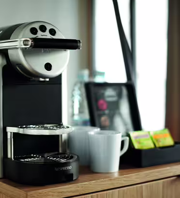 En moderne espressomaskine på en trædisk med to hvide krus ved siden af. I nærheden rummer en sort bakke et udvalg af teposer, sukkerpakker og en flaske vand. Baggrunden har et sløret vinduesgardin.