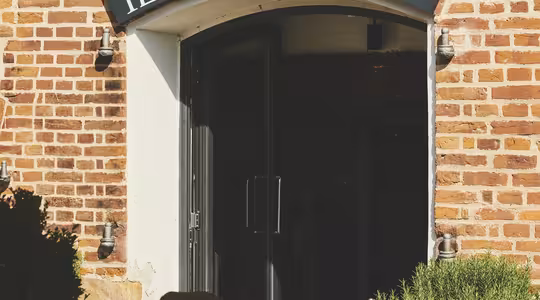 Indgang til "Il Rosmarino" restaurant med et ydre i mursten. Udendørs siddepladser er sat med borde og stole under en baldakin. Planter er ved siden af indgangen, hvilket skaber en indbydende atmosfære.